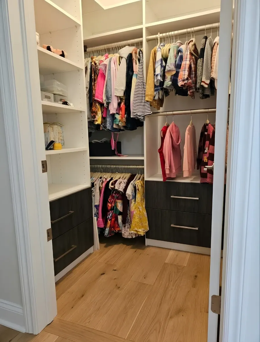 A small white walk-in closet for a child with two levels of hanging, adjustable shelves holding bins on the left, and a dark wood-grain drawer bank at the base, on an oak floor