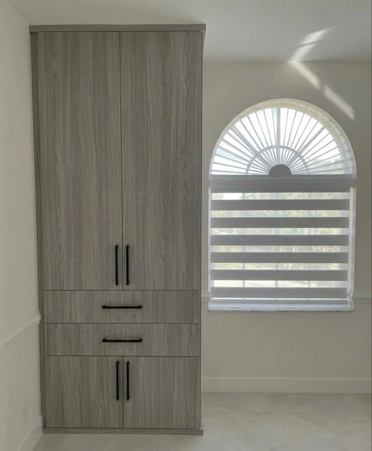 Tall grey woodgrain built-in wardrobe fitted into an alcove beside an arched window, with black bar pulls and drawers at waist height