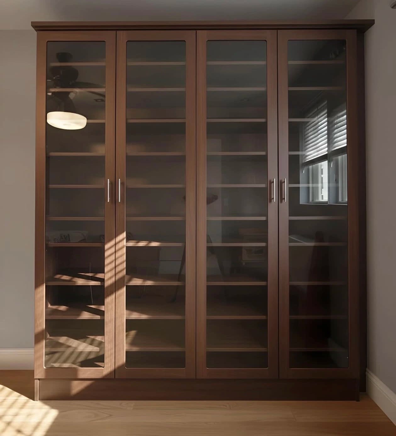 Dark walnut built-in wardrobe with four full-height glass doors over adjustable shelving, in a bedroom with afternoon light across it