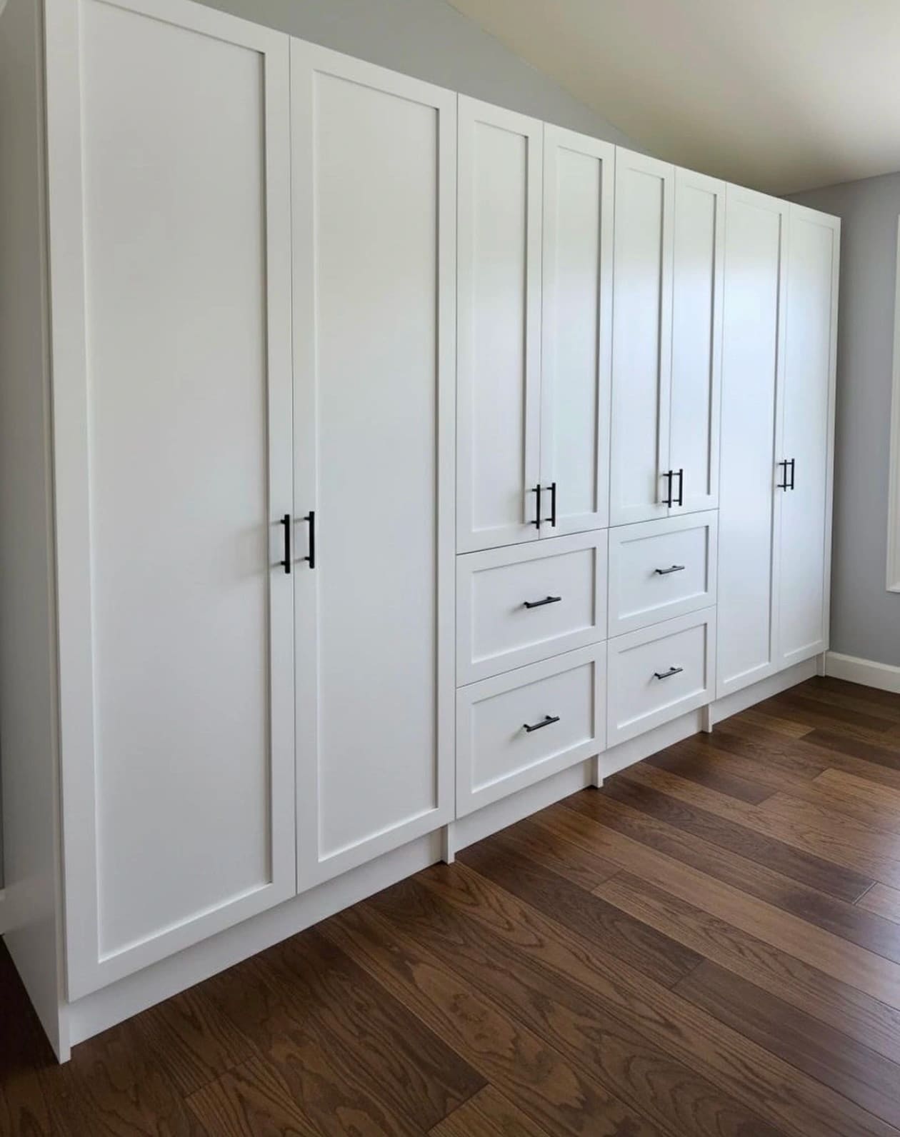 White shaker built-in wardrobe running the length of a bedroom wall, its top angled to follow the sloped ceiling, with a drawer bank in the middle