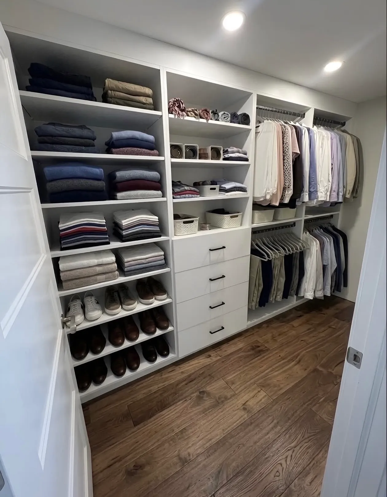 A white walk-in closet with a wall of folded shelving over shoe shelves, a drawer bank in the middle and double hanging along the far wall