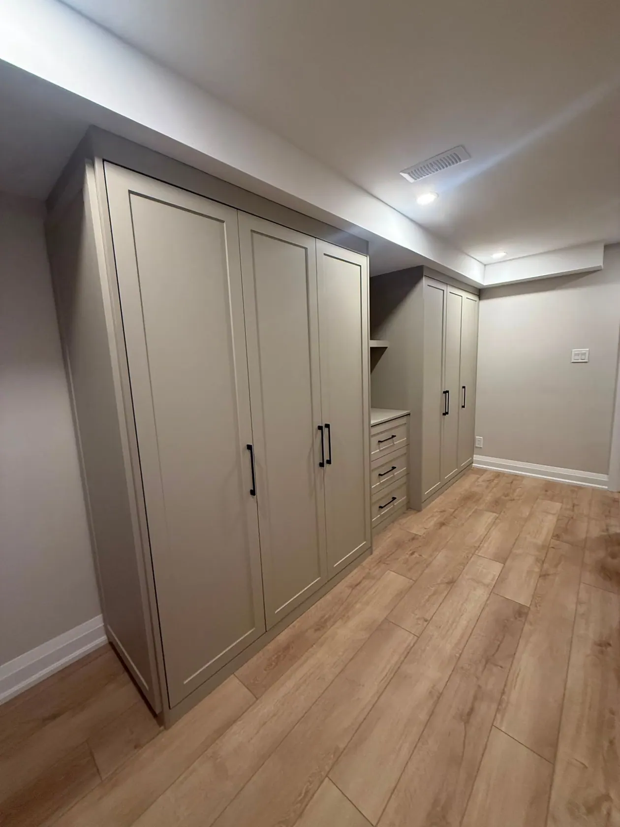 A run of greige shaker wardrobes with black handles beside a drawer bank and a second wardrobe bank in a basement bedroom