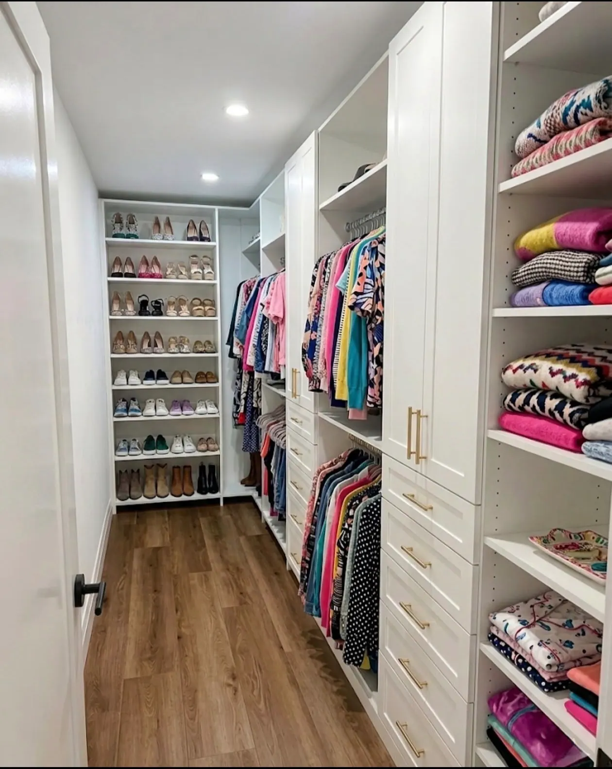 A white walk-in closet with a full-height shoe wall at the far end, hanging on the left and folded clothes on shelves to the right