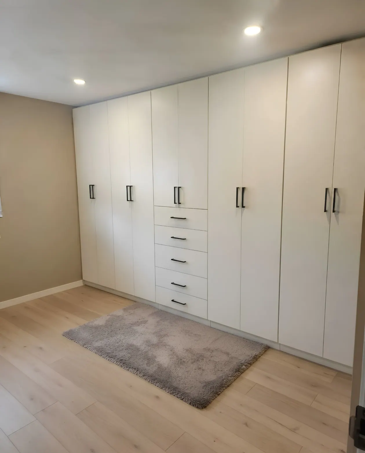 A wall of white flat-front wardrobes to the ceiling with black handles and a five-drawer bank in the middle of the run