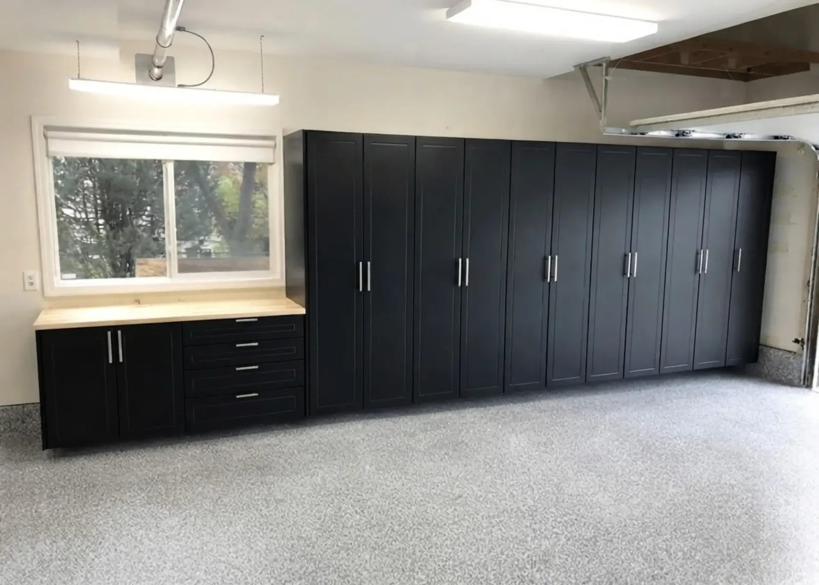 Black garage cabinets running the length of a garage wall, with a butcher-block workbench and four drawers under a window at the near end