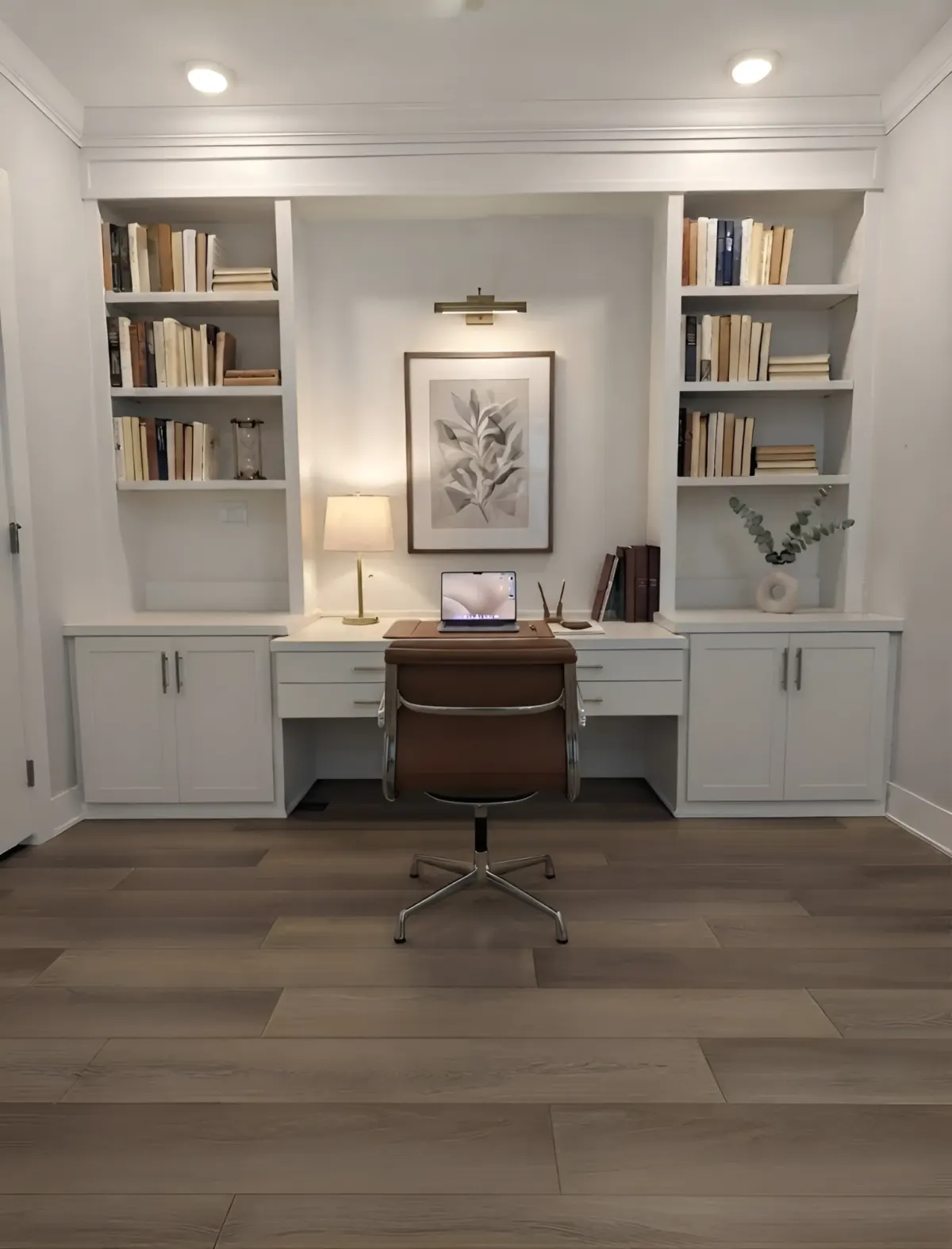 A white built-in home office - bookshelves either side of a framed print under a picture light, with a desk and drawer banks between them