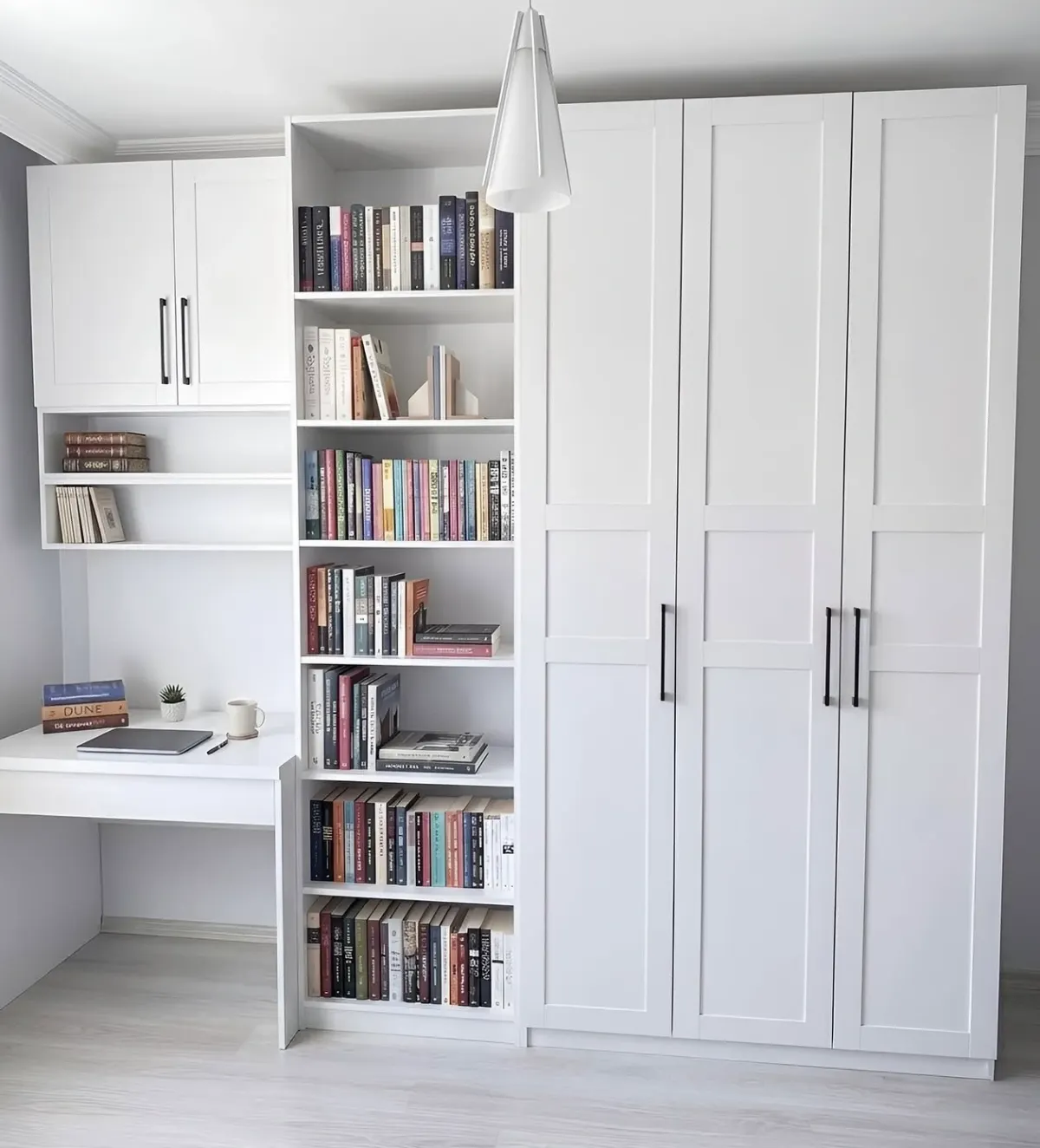 A white home office in a bedroom - a floor-to-ceiling bookcase full of books beside full-height wardrobe doors, with a desk and open shelves returning along the wall