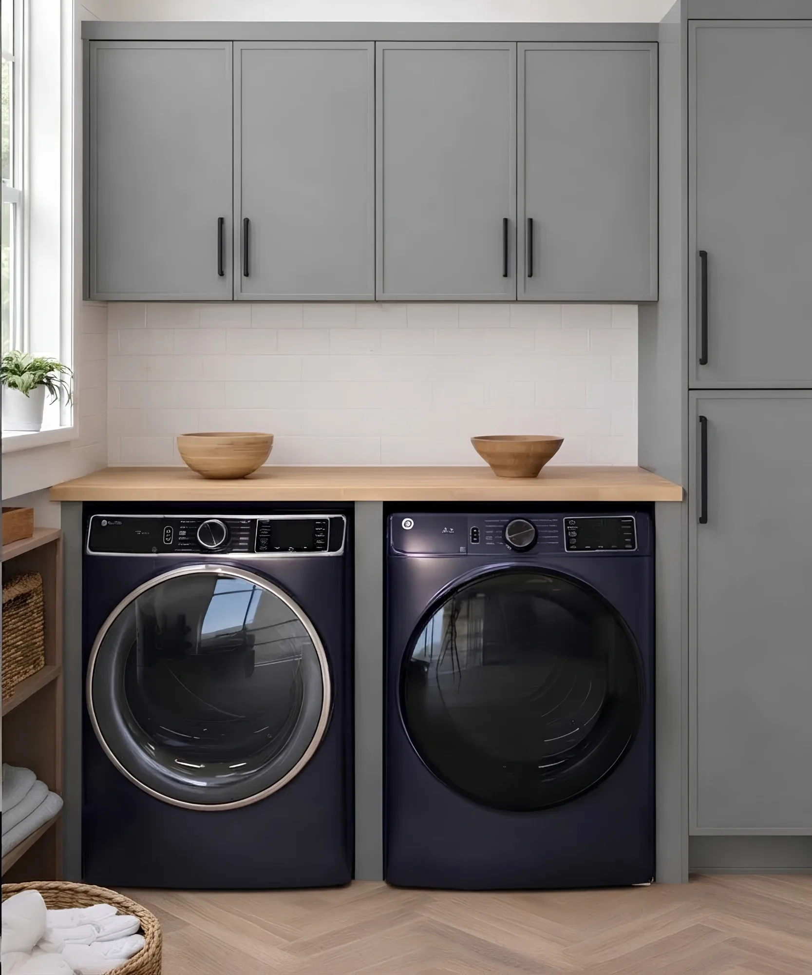 A grey laundry run: flat-front wall cabinets over a butcher block counter carried across two navy front-loaders, a full-height cabinet to one side and towels on wooden pegs