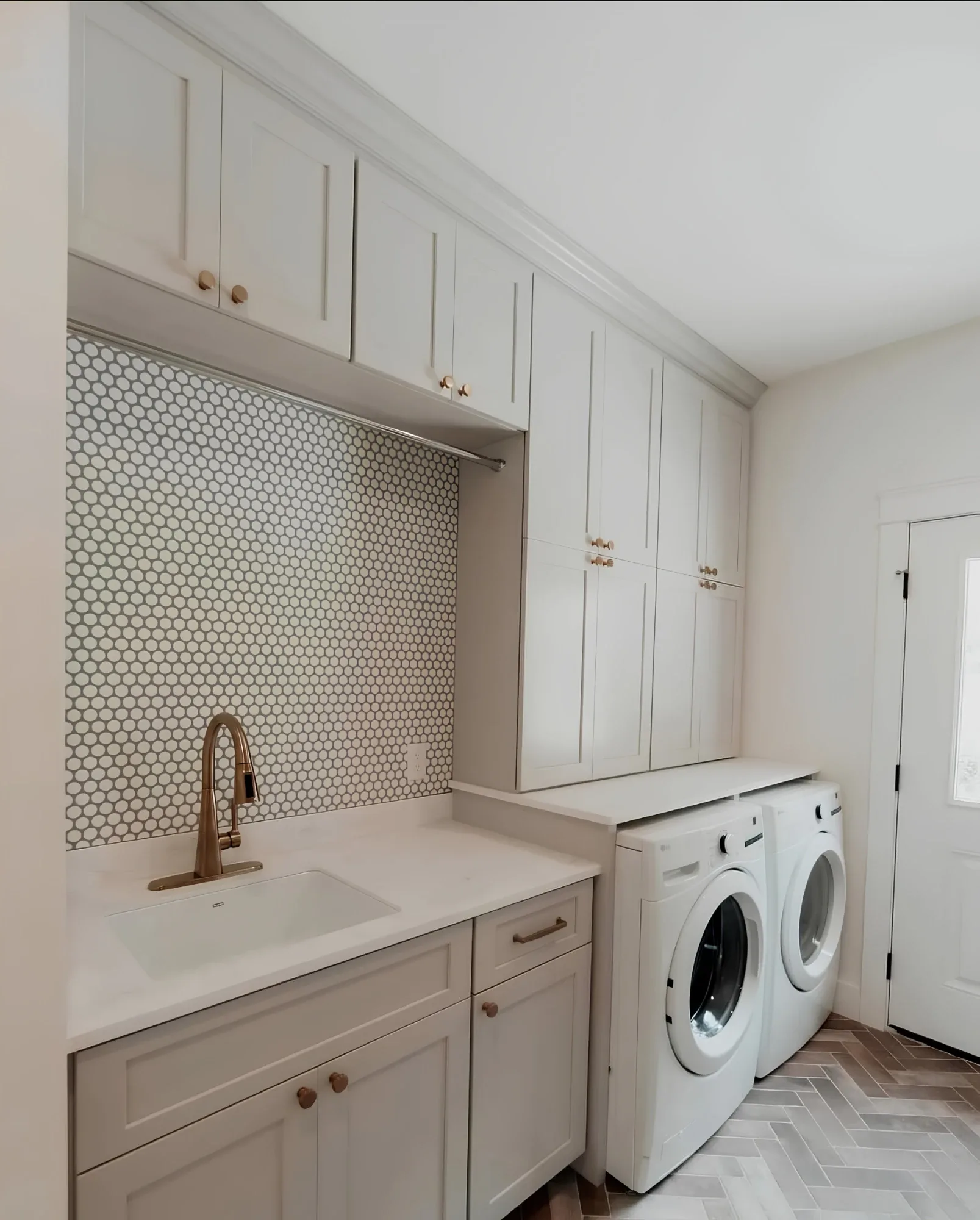 A cream laundry room with a penny-round backsplash and brass tap, a hanging rail under a bridging cabinet and floor-to-ceiling storage beside the machines