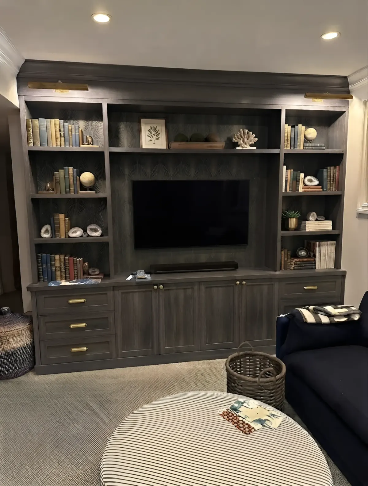 A dark oak media wall lit by two brass picture lights - bookshelves either side of a screen set against a patterned back panel, over drawers and cupboards