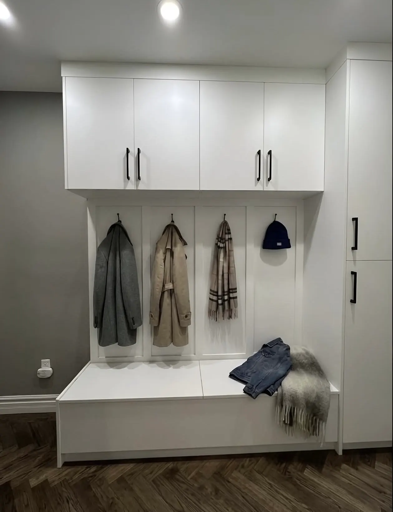 A white mudroom run with coats, a scarf and a hat on hooks above a bench, closed uppers over it and a tall cupboard alongside