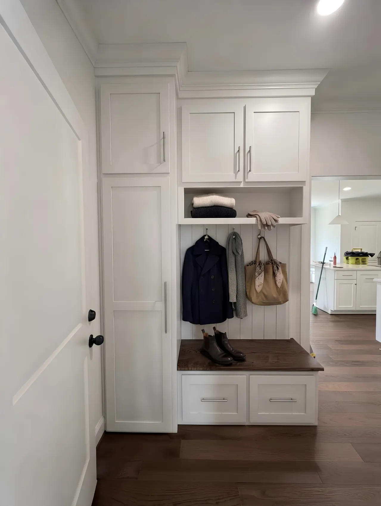 A white shaker mudroom by a door — a coat and bag on hooks, folded knitwear on the shelf above, and a dark timber bench over two drawers