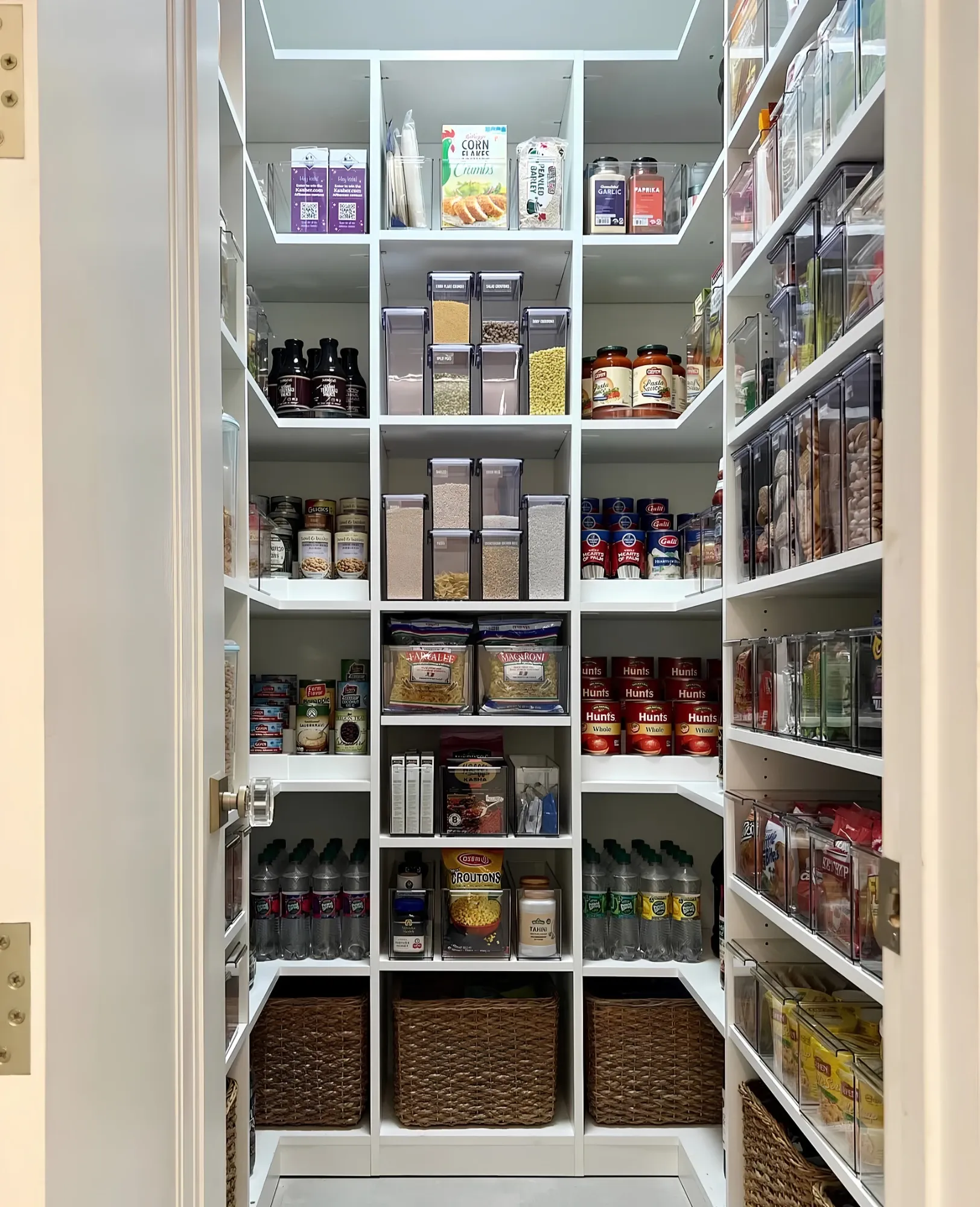 A white walk-in pantry shelved on three walls, clear stacking bins and canisters at eye level, tinned goods and bottles below and woven baskets on the floor shelf