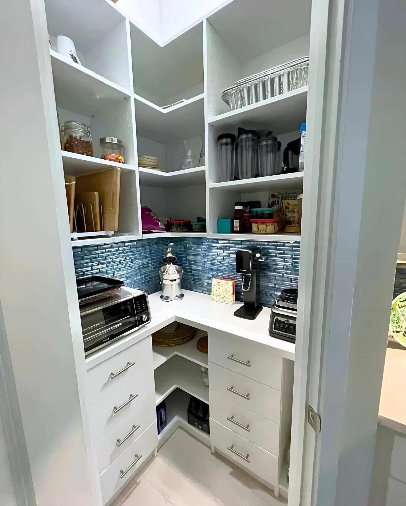 A white corner pantry with a blue glass tile backsplash over the counter, a toaster oven, juicer and blender on it, open shelving above and four-drawer banks either side