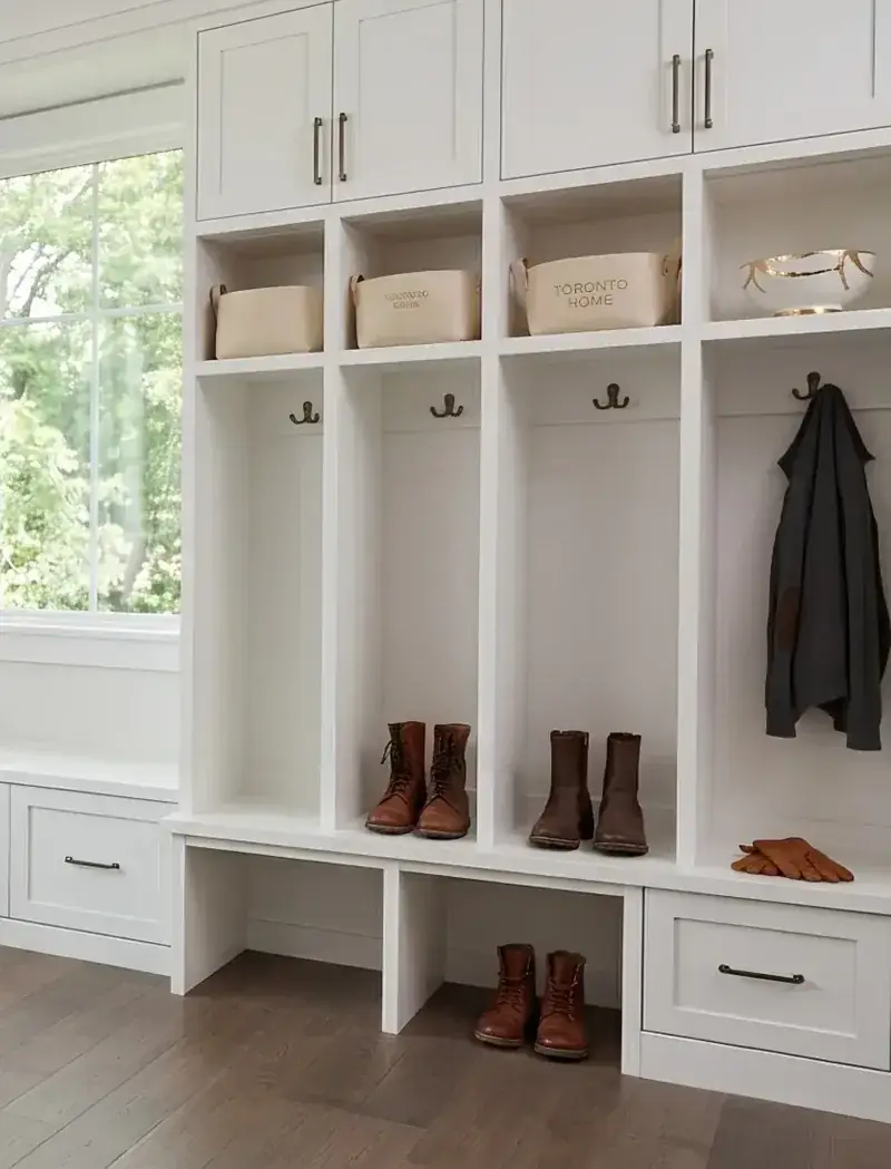 A white mudroom running two walls — a cushioned window bench with baskets under it, tall cupboards, and a run of lockers with hooks, boots and a coat
